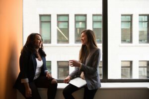 Two women in business outfits sitting by a window discussing something in a positive manner. You will benefit too.