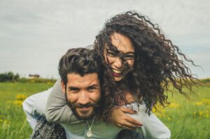 A man smiling and holding a woman on his back in the middle of a field while she laughs. Step into a new you.