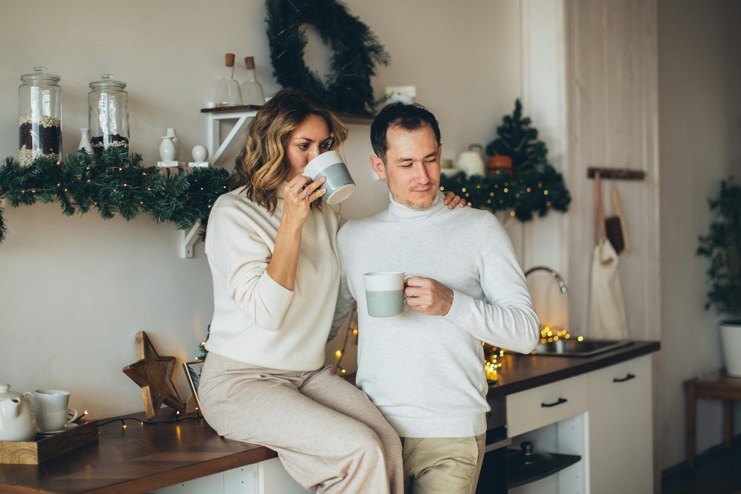 couple sitting and leaning on each other against the kitchen counter while drinking out of mugs