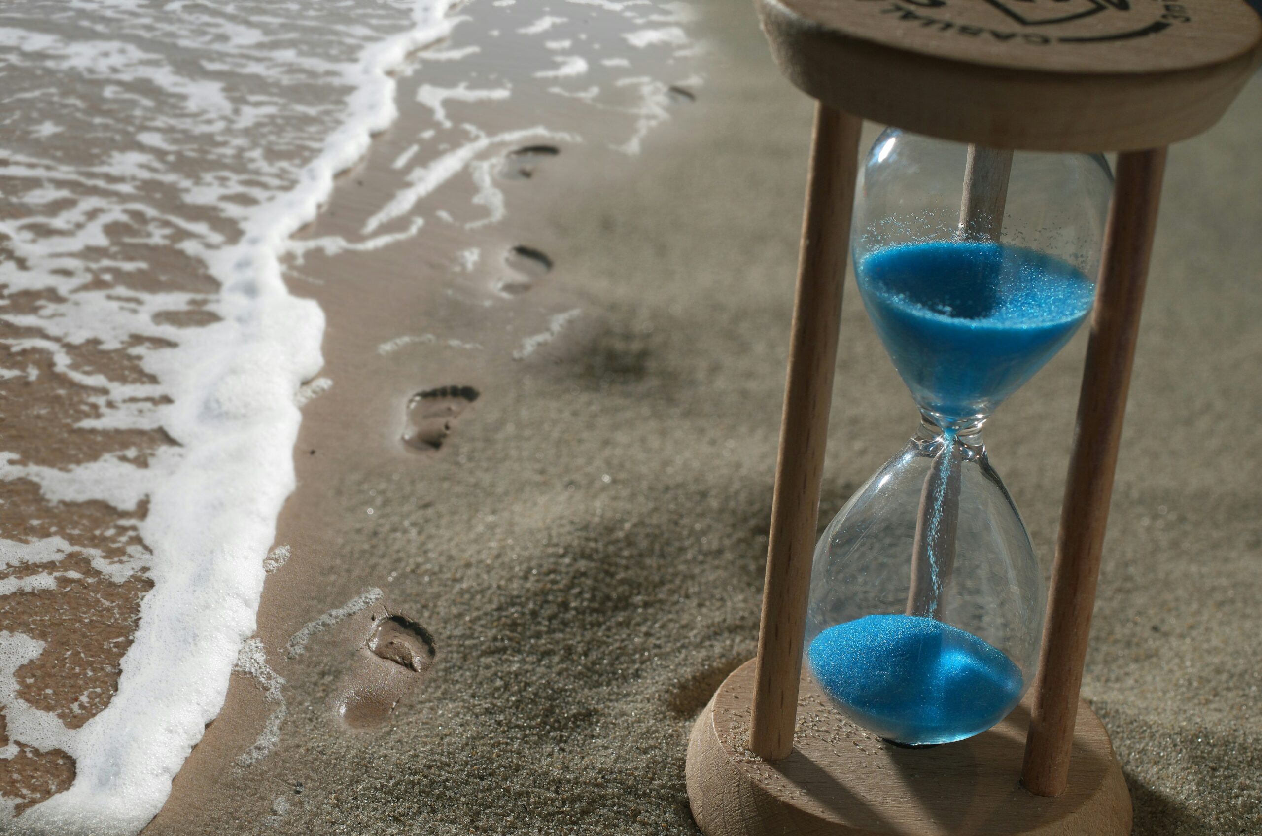 hourglass placed on the beach in the sand next to a set of footprints and gentle waves
