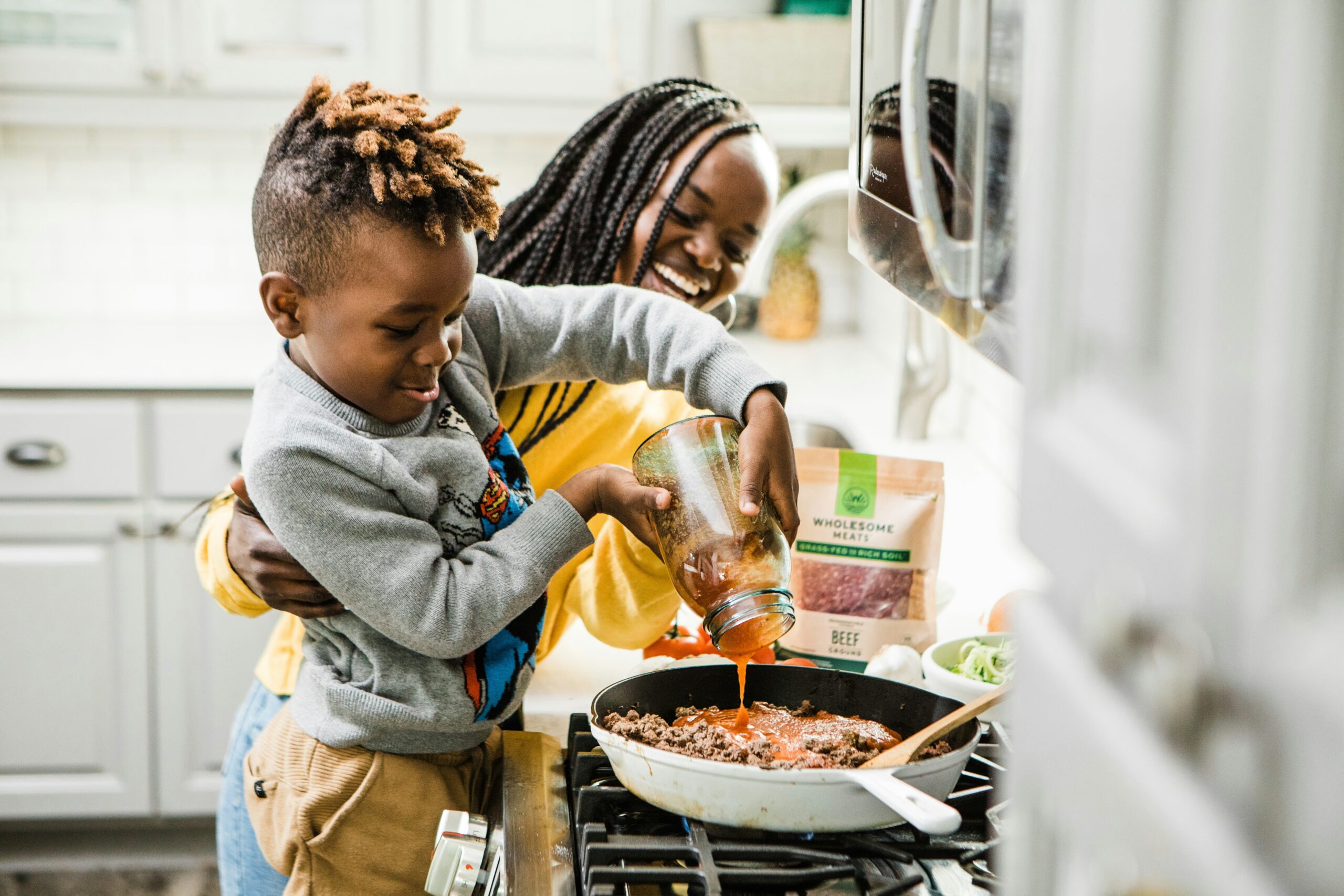 young boy pouring pasta sauce into sauce pan on stove top with the help of his mother, understanding and healing attachment impact