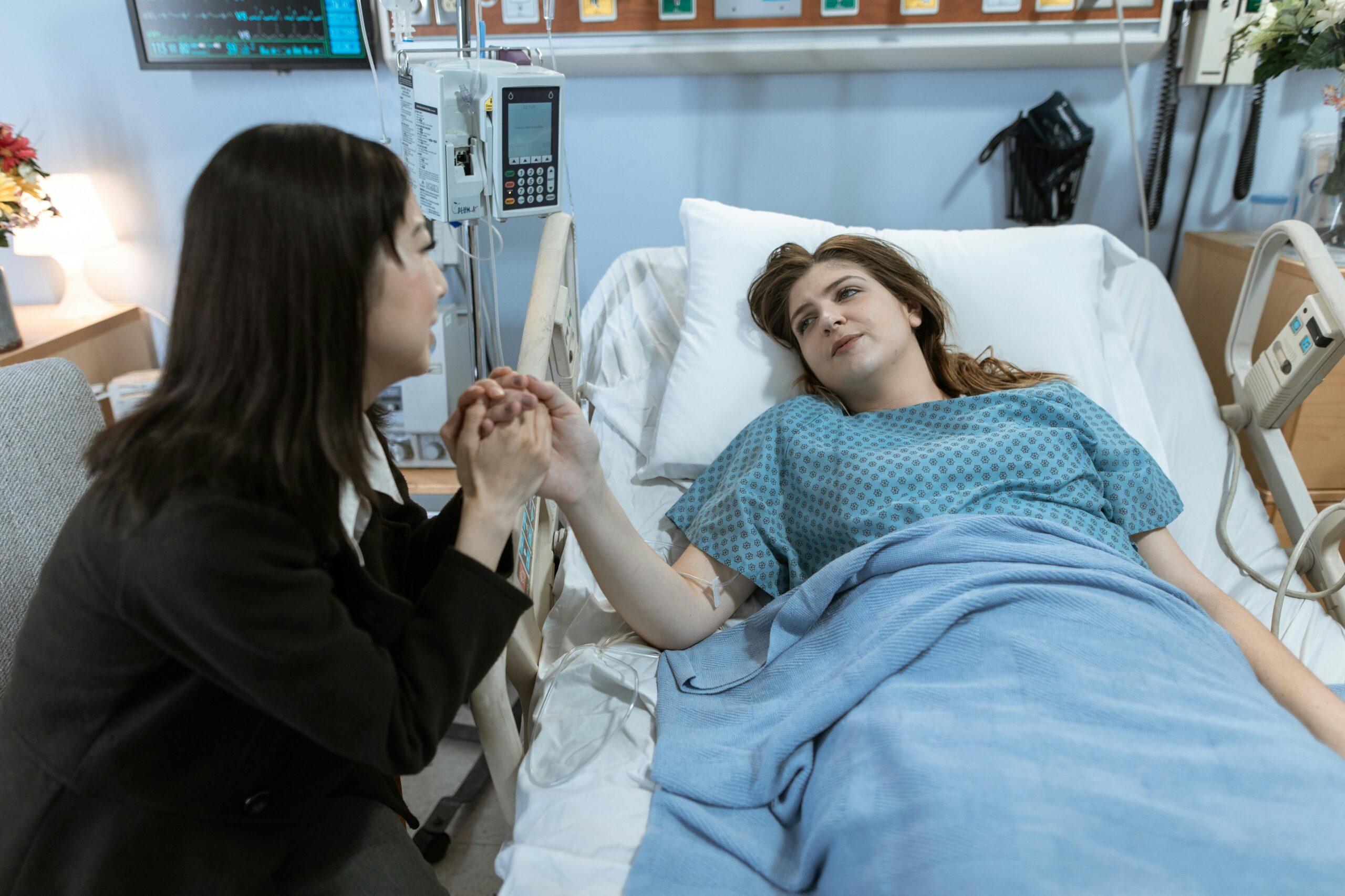 woman lies in a hospital bed holding hands with a visitor who sits beside her. The Key to Happiness & Health